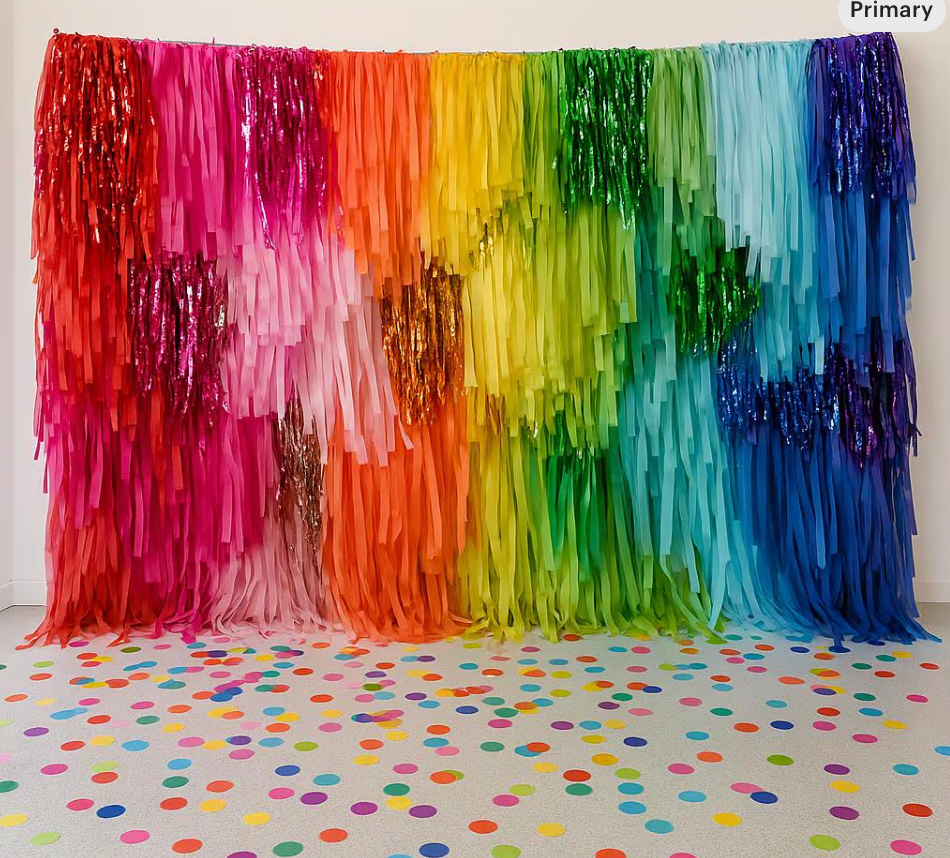 A colorful rainbow fringe streamer backdrop with a gradient of yellow, pink, orange, green, blue, and purple hanging on a wall with a dotted floor in the foreground.