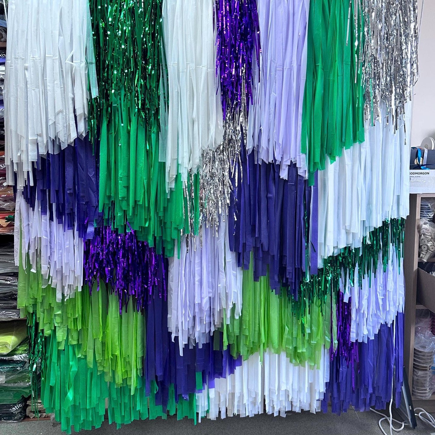 A handmade fringe backdrop featuring streams of purple, green, white, and silver hanging from the ceiling, designed for use in parties and events.